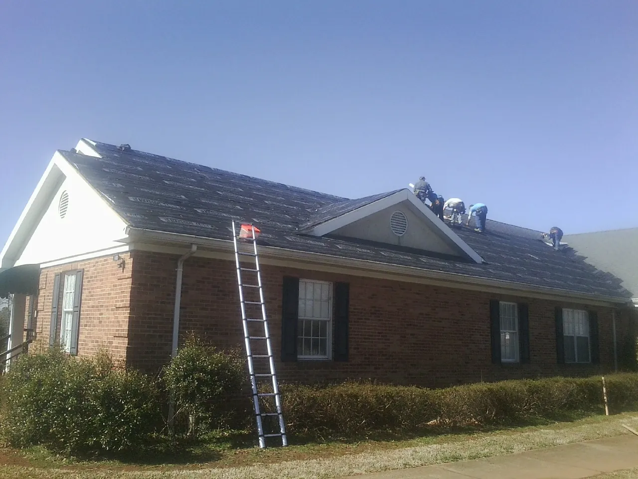 Crew tearing shingles off a brick ranch roof with a ladder set — Metal Roof Repair work by our crew in Belmont