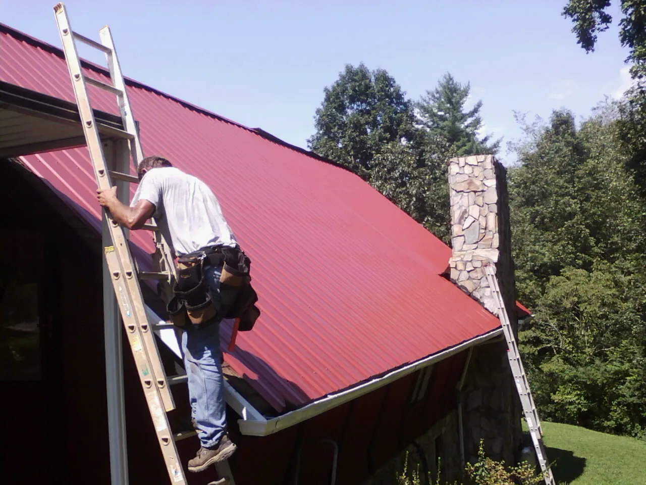 Roofer on a ladder beside a red metal roof and stone chimney — Soffit & Fascia Repair by Roofing Brothers in Belmont